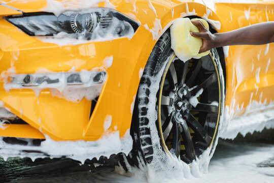 Hands of African man holding yellow sponge, washing car wheel with foam. Cleaning of modern rims of luxury yellow car at self car wash service outdoors.