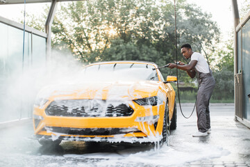 African stylish young focused man in work clothes washing his modern sport car with a water gun on...