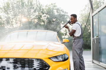 Handsome African young man in protective overalls, washing his yellow car manually with water high-pressure hose at outdoor self wash service. Car washing concept.