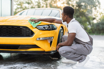 Young African man washing and wiping modern sport yellow car hood at the outdoor car wash self...