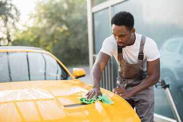 Car wash and cleaning at outdoors self service station. Shot of handsome bearded young African guy worker cleaning yellow car hood with a green microfiber cloth outdoors in summer sunny morning