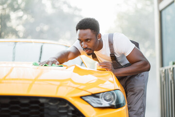 Handsome young casual African American man worker of self car wash service in gray overalls and white t-shirt wiping yellow car hood with green microfiber cloth at the open air car wash.