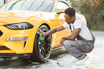 Naklejka premium Car wash service outdoors. Car wash self-service concept. Handsome African guy wiping wheel of modern luxury car with green microfiber cloth during the washing process outdoors.