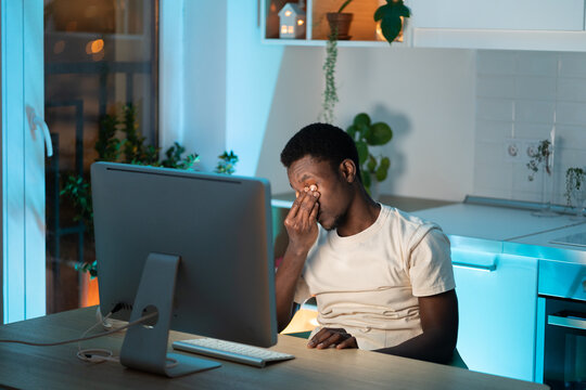Symptoms of overworking. Young tired overworked african man freelancer working late from home, sitting in front of monitor, massaging nose bridge, feeling eye strain and fatigue during computer work
