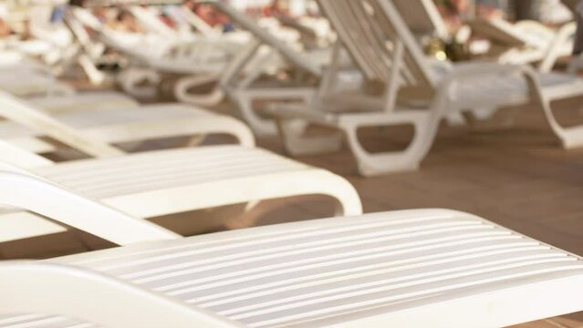 Empty white plastic sunbeds at holiday resort near pool, closeup detail, blurred people moving in background