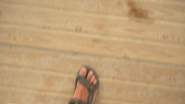 Looking down to young man feet in brown leather sandals walking over wooden boards path on sandy beach