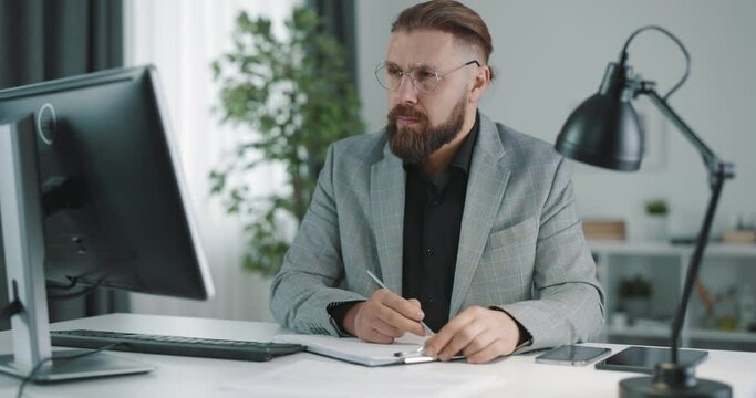 Focused bearded businessman writing notes while working on modern pac at bright office. Caucasian mature man wearing eyeglasses and stylish formal suit.