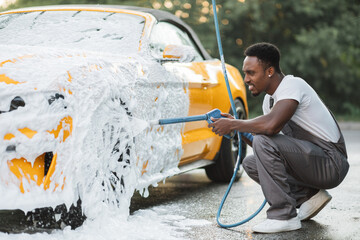 Young African man in gray overalls and white t-shirt washing wheels of his luxury car at self car wash station outdoors, spraying the foam with high pressure . Car wash outdoors concept.