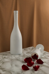 Minimal vintage style concept with white wine bottle, glass and red rose petals on marble table. Beige curtain background. Retro Valentine's day party composition.
