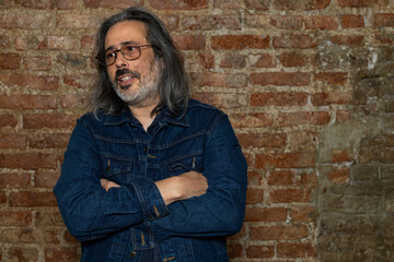 Latin American man (44) with long gray hair, blue jeans jacket and crossed arms. Copy space. Brick background. Lifestyle concept