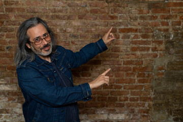 Latin American man (44) with long gray hair, blue jeans jacket points with both arms to his left side. Copy space. Brick background. Lifestyle concept