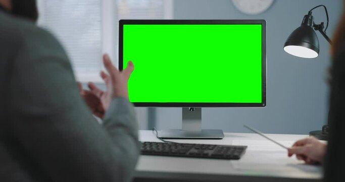 Back View Of Two Business Partners Looking Computer With Green Mock Up Screen While Sitting Together At Office Desk. Man And Woman Talking And Gesturing During Working Meeting.