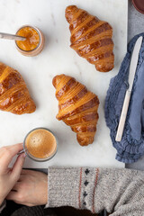 A relaxed white woman with a cup of foamy coffee in her hand next to three shiny delicious croissants.