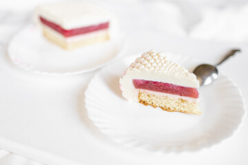 A white-pink piece of cake lies on a white plate on a light background. Delicious pastries