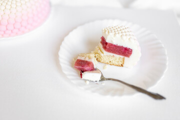 A white-pink piece of cake lies on a white plate on a light background. Delicious pastries