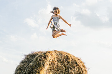 Naklejka na ściany i meble Portrait of little girl in dress playing and jumping on haystack in field. Light bright sunny day. Cheerful and enjoy freedom concept. High bounce. Reach sky. Side view. Low angle. One kid in field