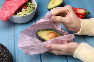 Woman packing half of fresh avocado into beeswax food wrap at light blue wooden table, closeup