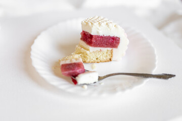 A white-pink piece of cake lies on a white plate on a light background. Delicious pastries
