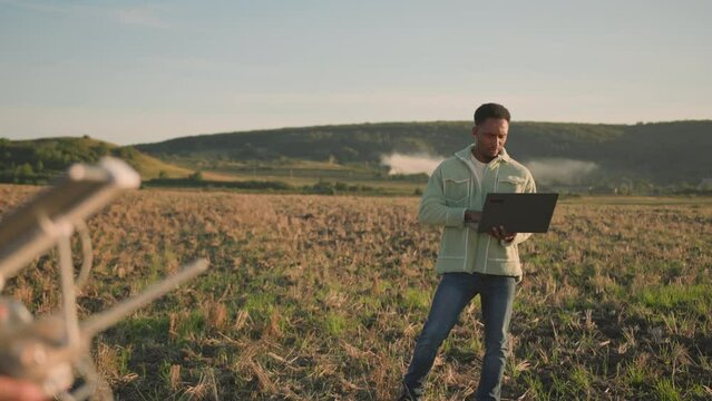 African farming specialist using computer laptop in the field. His colleague asian engineer flying drone with digital tablet. Cooperation. Agribusiness. Technologies.