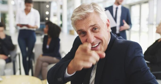Mature businessman in an office with his colleagues in the background. Mature caucasian businessman smiling happy talking and pointing his finger making a gesture sitting at work.