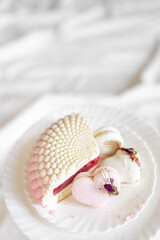 A white-pink piece of cake lies on a white plate on a light background. A pink heart-shaped cake lies on a white plate on a light background. Light macaroons on a white canvas. Delicious pastries