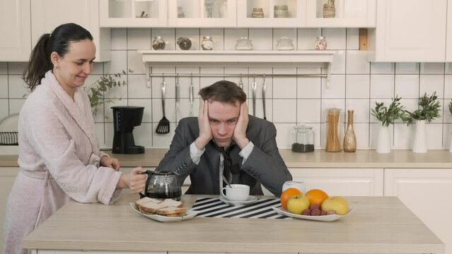 Young Woman Bringing Breakfast to her Husband. Exhausted Man Drinking from Coffee Pot while Sitting at Table in the Kitchen at Home. Business, Lifestyle, Overworking, Caffeine Addiction Concept
