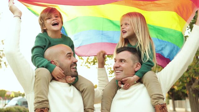 Gay male couple with children having fun holding LGBT rainbow flag outdoor at city park - Family and love concept