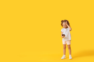 Fototapeta na wymiar Surprised little girl with mobile phone on yellow background