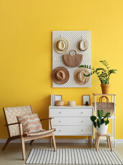 Stylish chair, chest of drawers, pegboard with hats and bag hanging on yellow wall