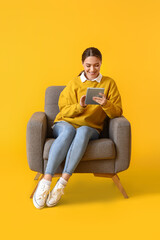 Pretty young woman using tablet computer in soft armchair on yellow background