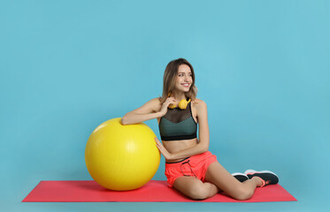 Fototapeta na wymiar Beautiful woman with headphones sitting on yoga mat near fitness ball against turquoise background