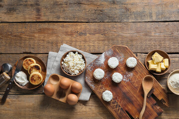 Board with uncooked cottage cheese pancakes and ingredients on wooden background