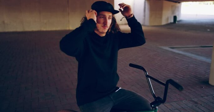 His style speaks for him. BMX biker adjusting his hat while sitting on his bicycle under a bridge alone outside. Confident cool young man with long hair wearing a cap and getting ready to do tricks
