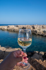 Woman's hand holding glass of white dry white wine with view on rocks and blue sea bay water near Protaras touristic town on Cyprus