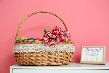 Fototapeta na wymiar Wicker basket with Easter eggs and flowers on table near color wall