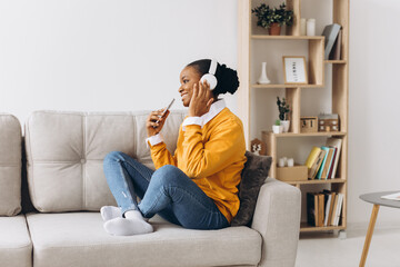 People, technology and leisure concept - happy african american young woman sitting on sofa with smartphone and headphones listening to music at home