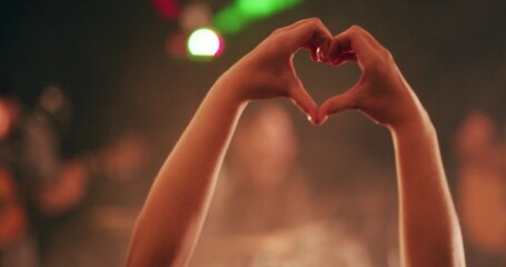Good music does the heart good. Hands of woman making a heart shape love gesture during a live music performance. Audience having fun and enjoying a festival or party vibe with strobe lights