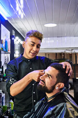 barbero joven cortando el pelo de uno de sus clientes, mientras lo atiende con una gran sonrisa y...