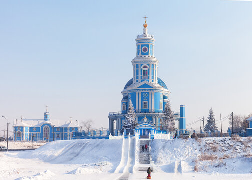 The beautiful blue bell tower of the Kazan Orthodox Church on a hill in the village of Telma, Irkutsk region. Children sledding down an ice slide during Christmas holidays on a cold winter day
