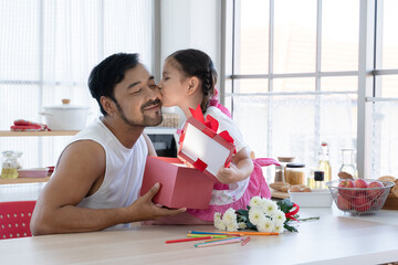 Little mixed race daughter kissing young Asian dad on cheek while giving and opening gift box together. Family sitting at home kitchen celebrating father day or birthday special occasion