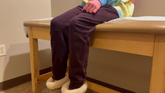 Young kid child girl or boy in covid-19 mask at Doctor&rsquo;s office hospital visit waiting on examination table nervously fidgeting kicking feet semi-close shot panning down on Ugg slipper shoes feet.