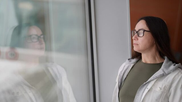 calm pretty adult woman with glasses for vision is standing near large window, portrait of lady