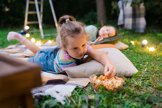 Family summer outdoor movie night. Girl lying on blanket and  pillows, eating homemade popcorn and watching film on  DIY  screen with from projector. Summer outdoor weekend activities with kids.