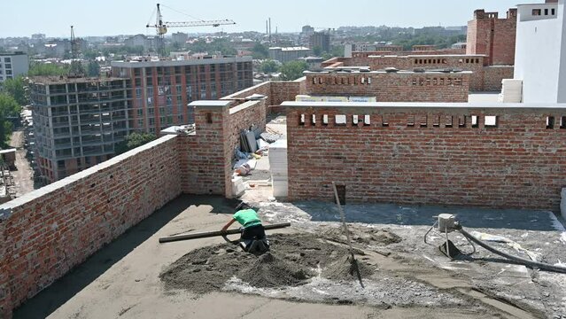 Construction site with newly built apartment buildings, tower crane and floor screeding contractor. Male worker using screed rail while screeding floor in building with brick walls.