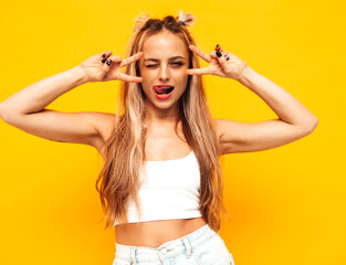 Young beautiful smiling blond female in trendy summer clothes. Sexy carefree woman posing near yellow wall in studio. Positive model having fun indoors. Cheerful and happy.Shows peace sign