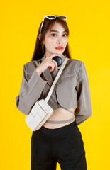 Portrait studio shot of Asian trendy fashionable female hipster teen model in casual crop top street wears jacket sunglasses carrying leather handbag purse standing look at camera on yellow background