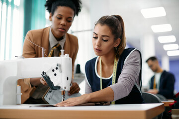 Young dressmaker works on sewing machine while being supervised by production line manager.