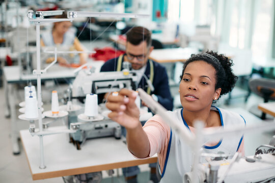 Young African American dressmaker adjusts sewing machine while working in clothing factory.