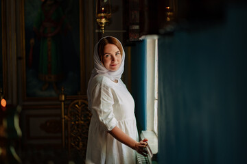 a woman in headscarf and light-colored robes by window in church. 