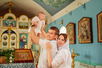 man and woman with baby son in light-colored robes by window in church. 